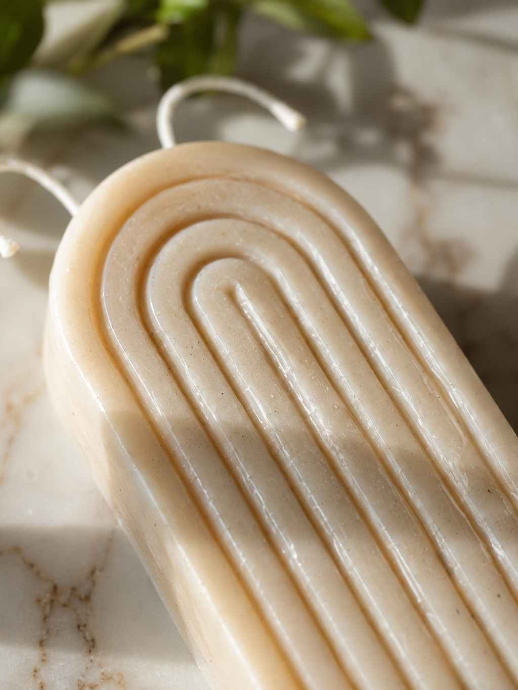 A close-up of a cream-colored soap bar with a ribbed, arch-like pattern, resting on a light marble surface with soft shadows and green leaves in the background.