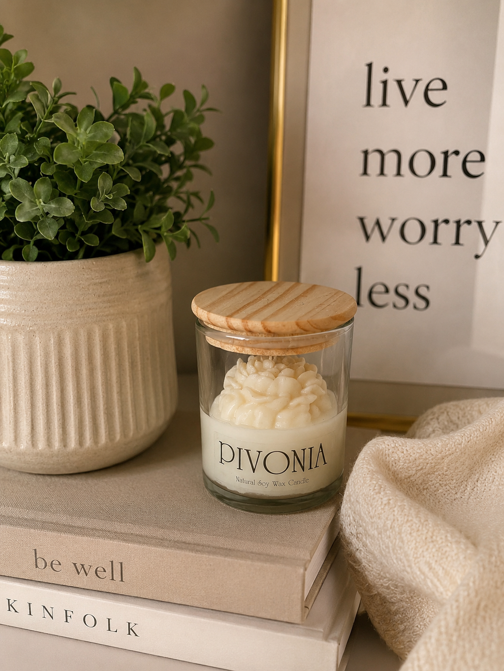 A glass jar candle with a light wood lid sits on stacked books beside a potted green plant and a cream-colored textured throw blanket. The candle label reads 'DIVONIA'.