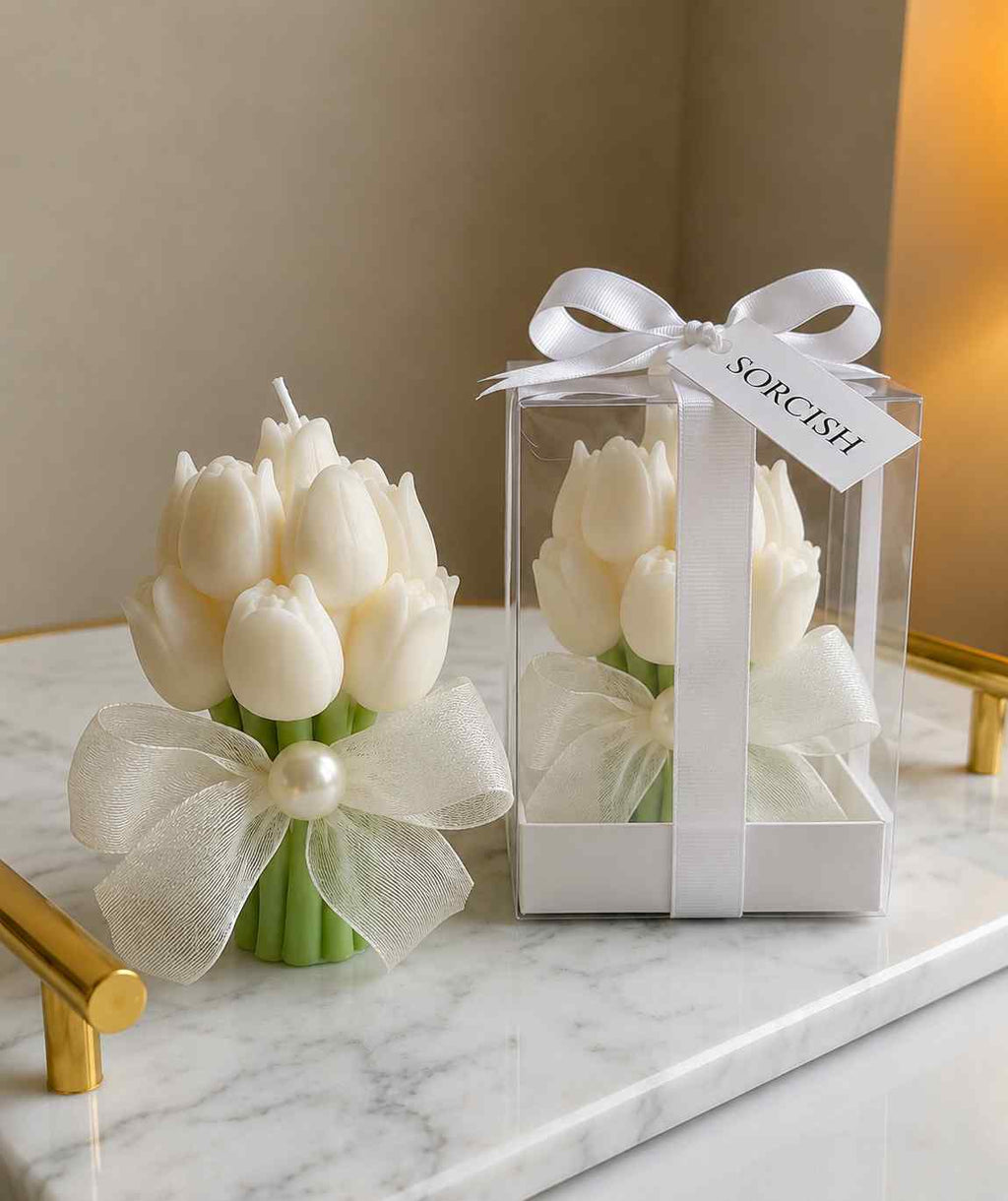 A bouquet-shaped candle made of white tulips with green stems and a pearl accent, shown next to its clear gift box with a white ribbon and bow. The box has a tag that reads 'SORCISH'.