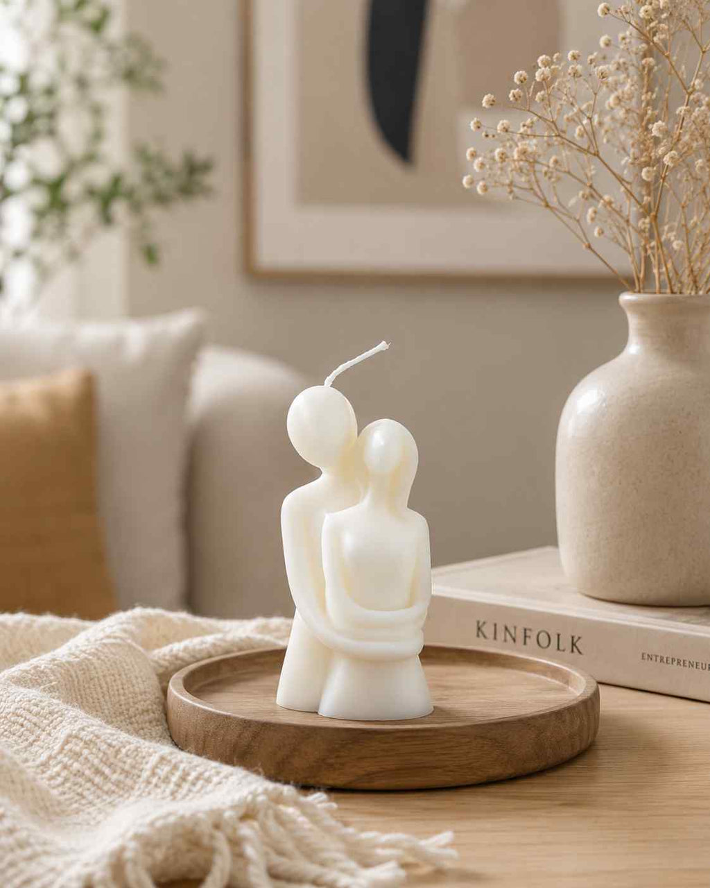 Candle shaped like a couple on a wooden tray with a vase and books in the background