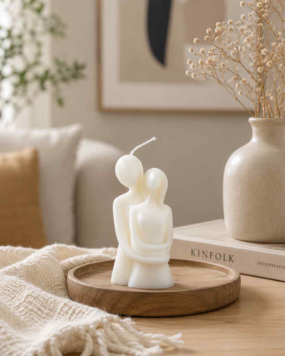 Candle shaped like a couple on a wooden tray with a vase and books in the background