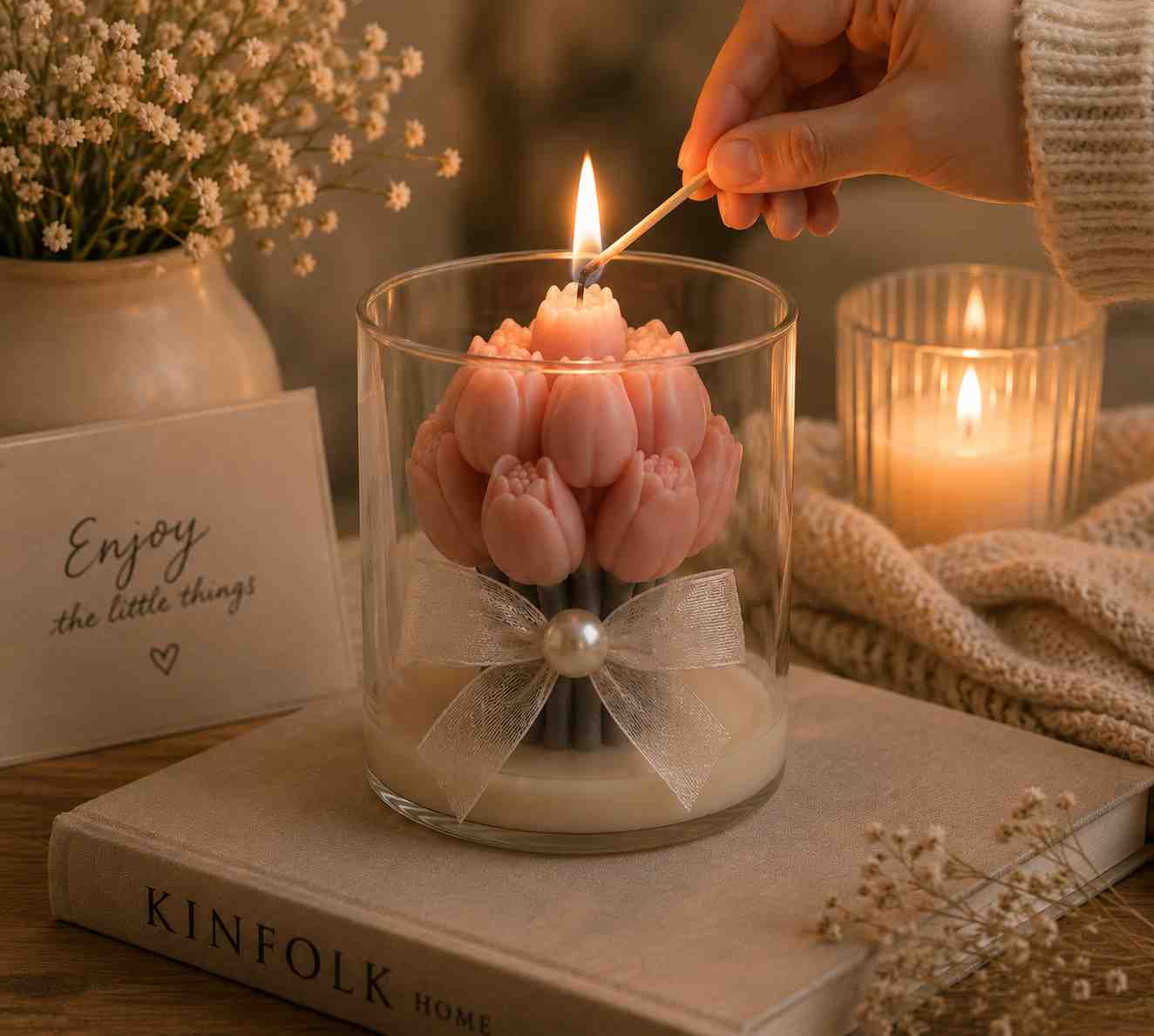 A pink tulip-shaped candle in a clear glass jar with a white ribbon bow, lit with a match held by a hand. The candle sits on a book on a wooden table with cozy background props including dried flowers, a knitted blanket, and another lit candle.