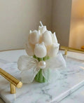 A bouquet-shaped candle made of white tulip shapes with green stems, tied with a sheer white ribbon and a pearl accent, displayed on a marble tray.