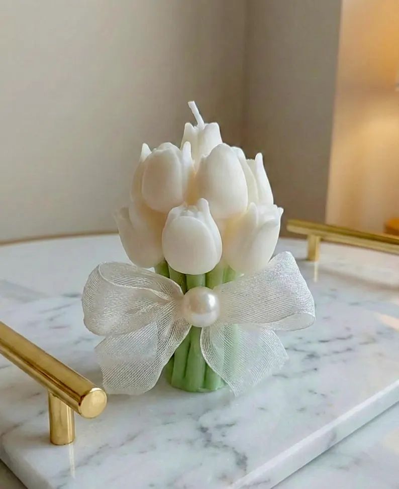 A bouquet-shaped candle made of white tulip shapes with green stems, tied with a sheer white ribbon and a pearl accent, displayed on a marble tray.