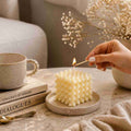 Hand lighting a textured candle with a cup, books, and dried flowers on a table.