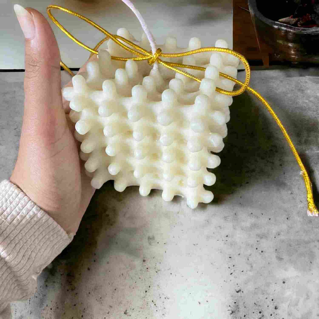 A hand holding a white, textured, coral-like wax candle with a yellow string and a white wick, set against a gray stone-like surface.