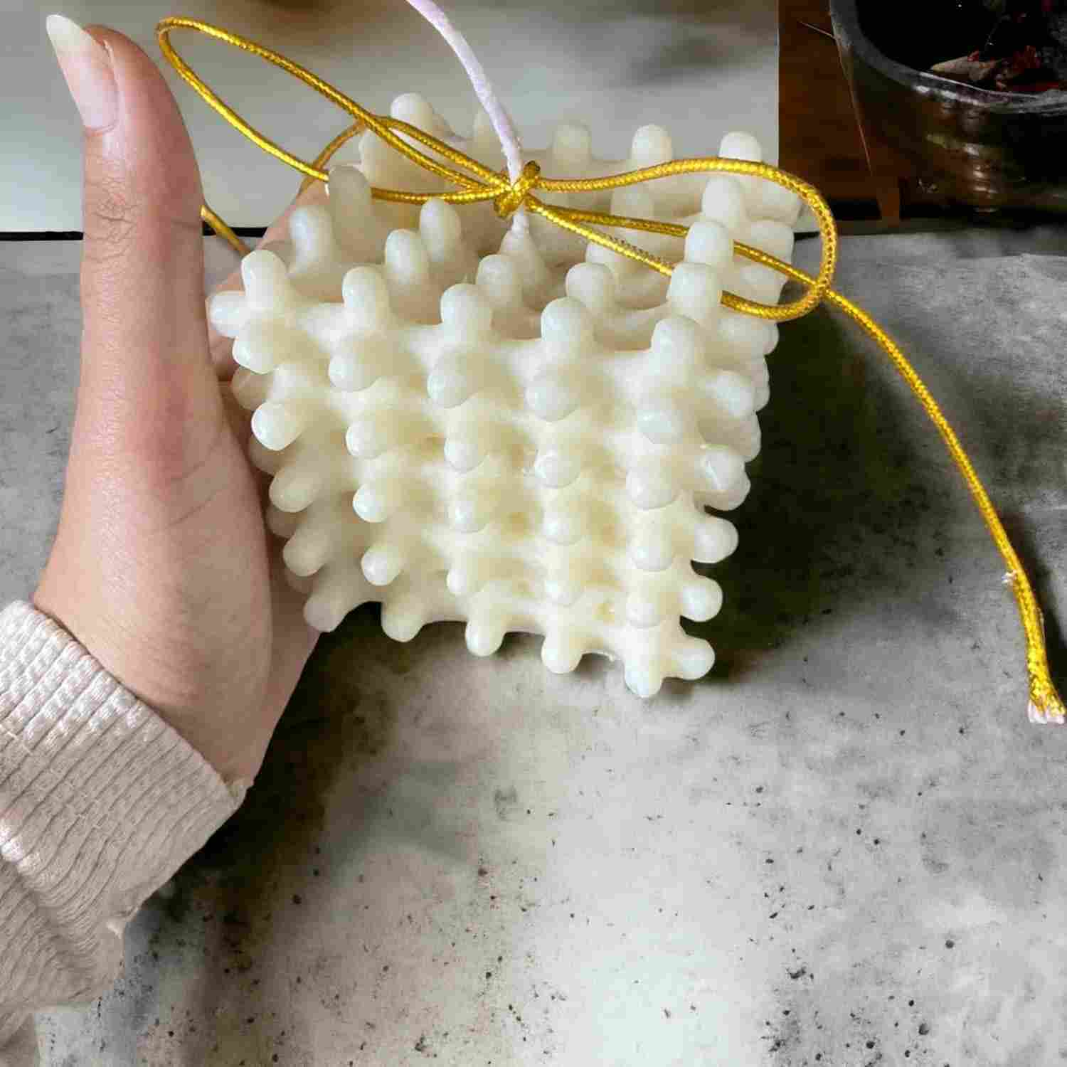 A hand holding a white, textured, coral-like wax candle with a yellow string and a white wick, set against a gray stone-like surface.