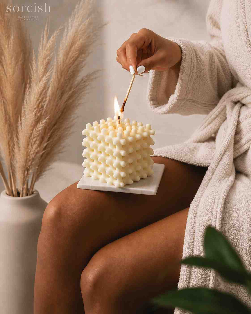 A person in a cream bathrobe sits beside a small white candle made of stacked pom-pom spheres, while a hand lights the candle with a wooden match. A white vase with dried pampas grass is on the left, and the scene is softly lit in a cozy indoor setting.