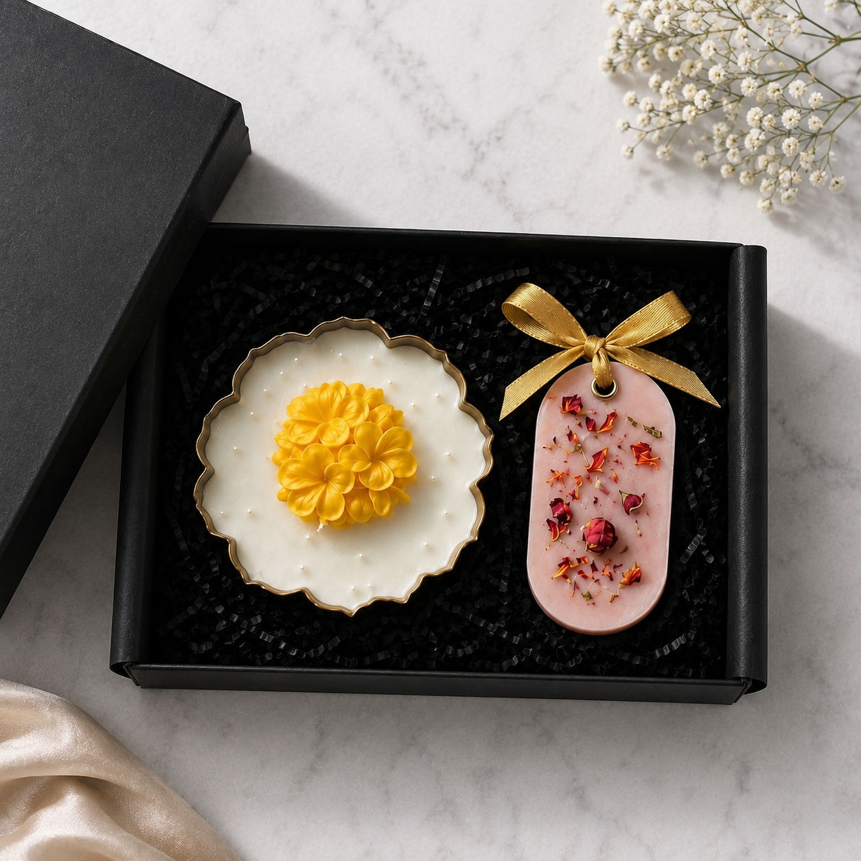A black gift box with two decorative items on a white marble surface: a white scalloped dish with a yellow flower-shaped center and a pink oval tag tied with a gold ribbon and decorated with dried red flowers. A black box lid sits beside the open box, with small white flowers in the top right corner.