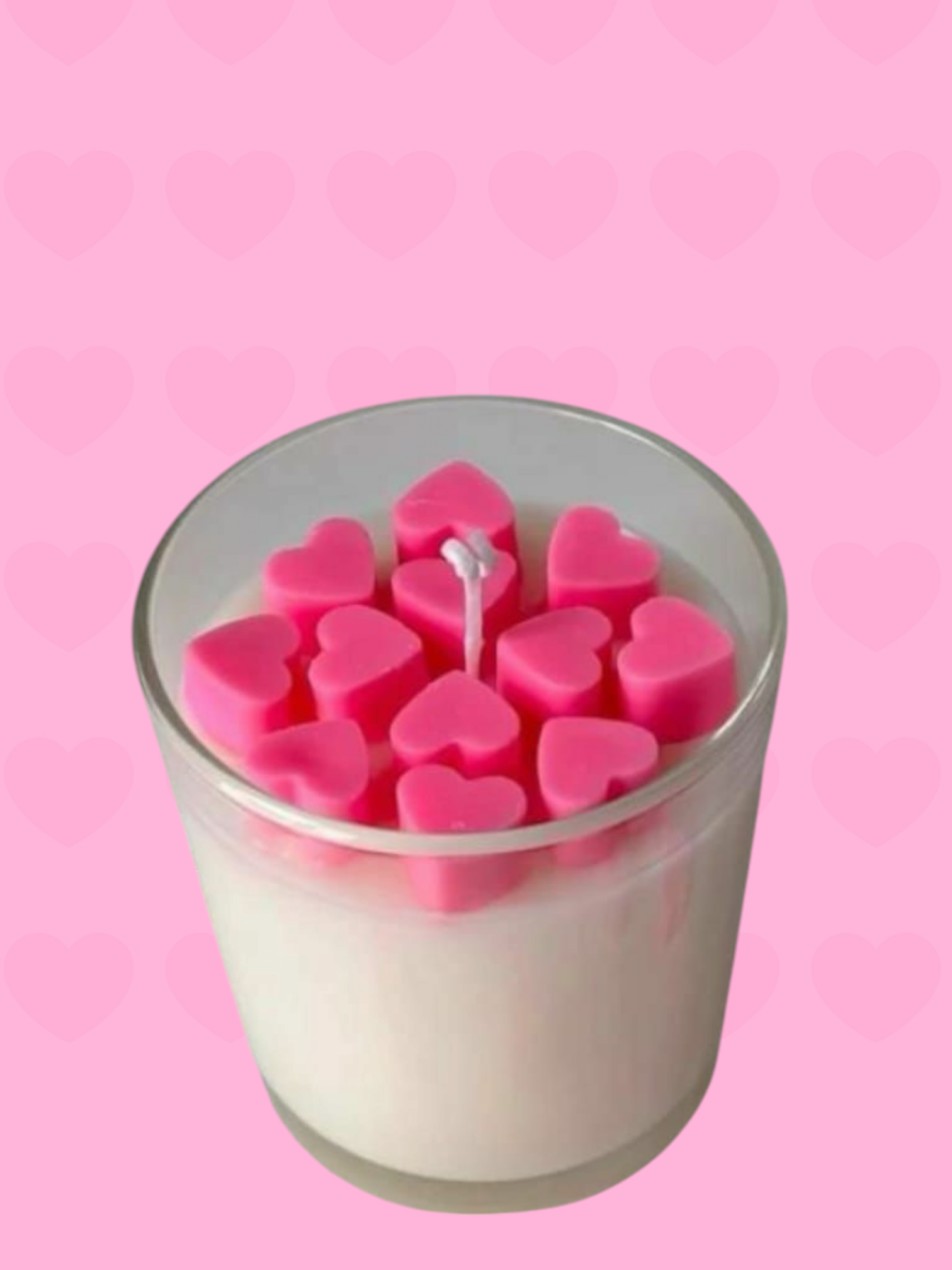 A transparent glass jar containing a creamy white soy wax candle with pink heart-shaped wax melts on top, set against a pink background.