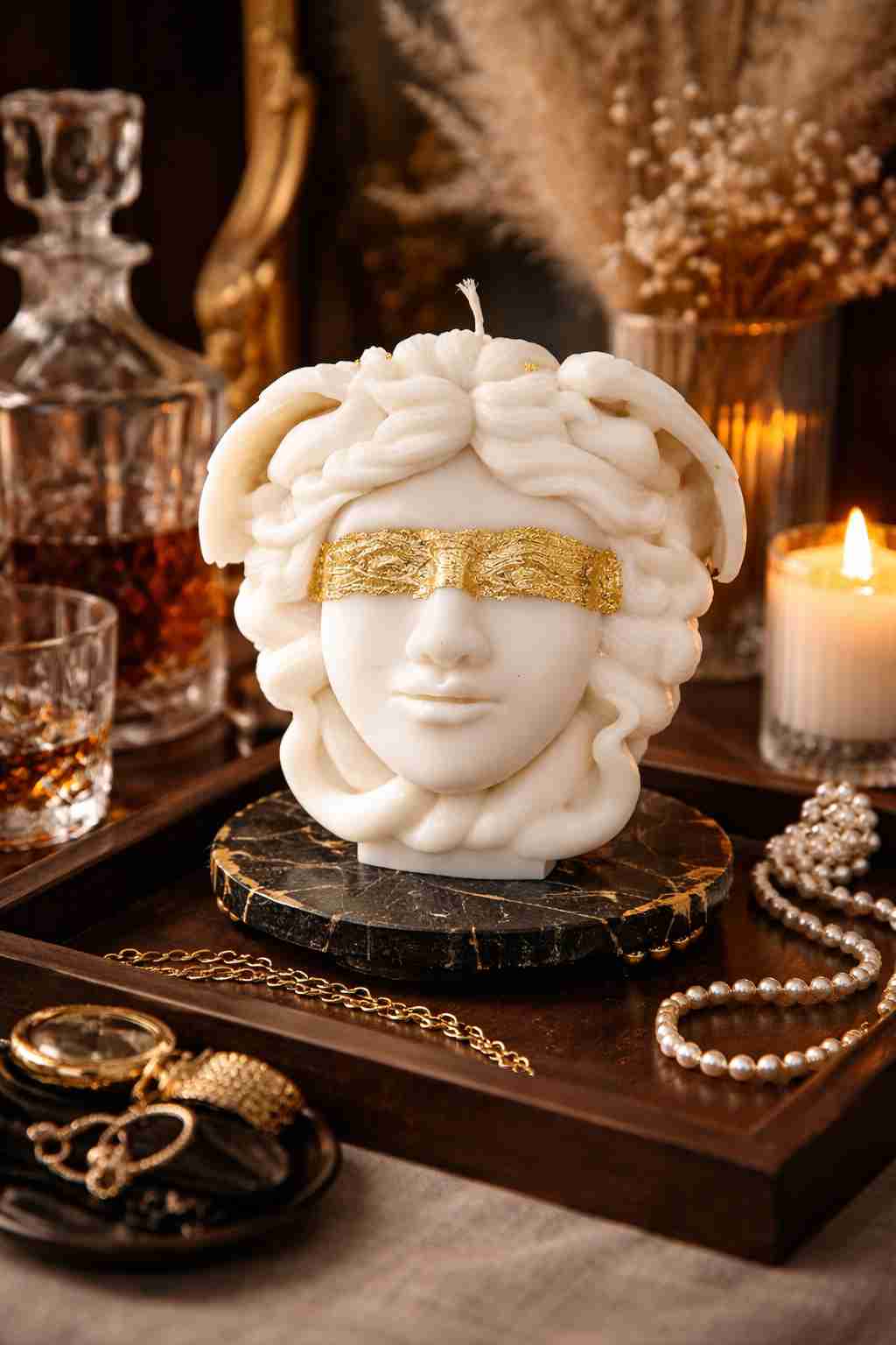A white sculptural candle shaped like a classical female head with wavy hair and a gold blindfold, displayed on a dark marble base on a wooden tray with candles, jewelry, and a decanter in a warm, moody setting.