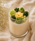 A glass jar candle filled with creamy off-white wax and decorated with sculpted green succulent and cactus candles, including a small yellow lotus flower in the center with a flame. The candle is set on a beige textured fabric background with dried white flowers on the left.