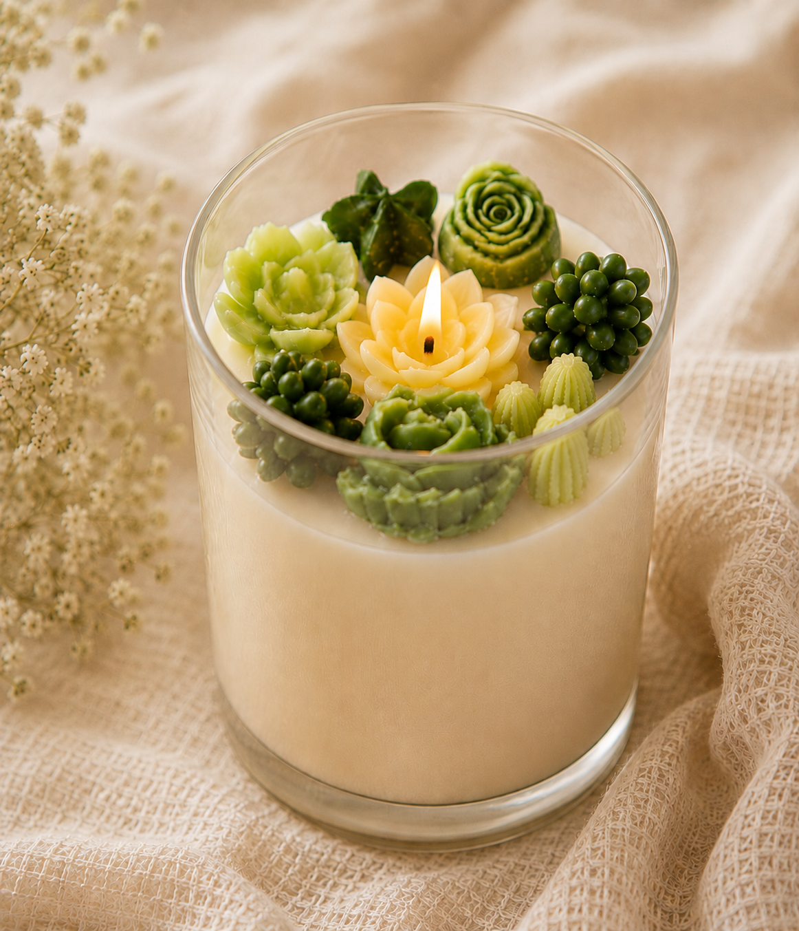 A glass jar candle filled with creamy off-white wax and decorated with sculpted green succulent and cactus candles, including a small yellow lotus flower in the center with a flame. The candle is set on a beige textured fabric background with dried white flowers on the left.