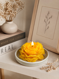 Yellow flower-shaped candle on a white plate with a vase of dried flowers and books in the background.