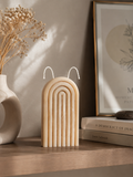 A beige arched candle with white wicks sits on a wooden shelf in a styled interior setting, next to a white ceramic vase with dried flowers and a framed print.